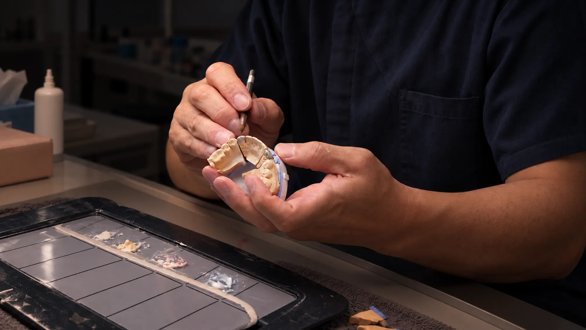 A master ceramist in navy scrubs at the bench, a fine brush in one hand and the case's stone model in the other